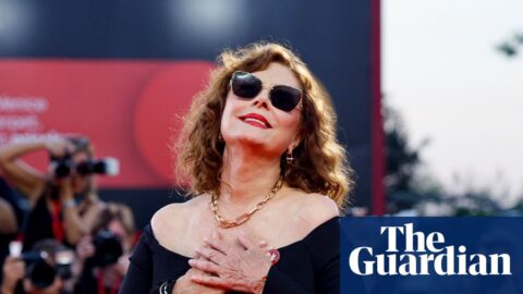 Susan Sarandon poses in sunglasses and an off-shoulder black dress as photographers shoot behind her