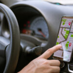A smartphone mounted beside a car's dashboard displays turn-by-turn navigation while a driver reaches toward the screen.